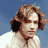 Pixelated static image of a person with light brown curly hair wearing dark earpieces and a white top against a blue background.