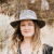 A pixelated static image shows a person with long light brown hair wearing a wide-brimmed dark hat with a light gray band, standing against a blurred natural background.