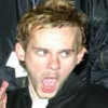 A close-up of a person with light brown hair wearing a dark jacket over a white shirt, making a surprised expression with mouth open and eyes wide.