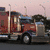 A red semi-truck with a blue cab is shown in against a pinkish sky background, with text on the right side