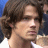 Pixelated close-up of a person with curly brown hair, light skin, and a neutral expression against a blue and white background.