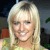 Pixelated close-up of a person with long blonde hair wearing a light green top, smiling with visible teeth against a dark background with blurred shapes.