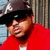 Pixelated image of a person wearing dark cap and red shirt, standing against vertical striped background with light blue section on left.