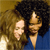 Two figures with dark curly hair, one in blue clothing, positioned closely together against a brown and blue background.