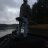 Pixelated grayscale image of a tall dark figure standing on rocky terrain near water with overcast sky background.