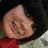 A person with short black hair and a red top smiles broadly, showing teeth, against a light background with metallic elements.