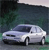 Pixelated image of a light gray sedan with dark tinted windows, positioned on a road against a blurred green and dark background.