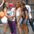 Group of people walking together, one wearing pink headband and purple pants, holding orange and white objects.