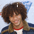A person with curly brown hair wears a dark blue jacket over a white shirt and red choker, smiling against a light background.