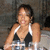 A woman in a black and white halter top smiles at the camera, seated with hands resting on a table holding two clear glasses.