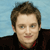 Pixelated portrait of a person with short brown hair, light skin, and a neutral expression wearing a dark jacket over a red-collared shirt against a light blue background.