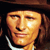 Pixelated close-up of a person's face with long brown hair, wearing a dark hat and white collar, against a dark background.
