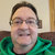 Pixelated portrait of a person with short dark hair wearing glasses and a green garment against a light background with a brown horizontal band.