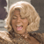Pixelated image of a person with blonde hair wearing a brown patterned top, smiling with mouth open showing teeth and tongue.