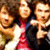 Three individuals in a group pose with blurred facial features and pixelated rendering, wearing dark outer garments over red and white clothing against a brown background.
