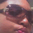 Pixelated close-up of a person with dark hair and light skin, wearing sunglasses and holding an object near their face.