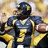 Pixelated image of a football player in dark blue uniform with yellow accents holding a brown football, standing against blurred background.