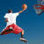 A basketball player in mid-air shooting a ball, wearing white shirt and red shorts against blue sky background.