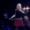 A woman with long blonde hair performs on a dark stage, wearing a black sleeveless top and red skirt while holding a microphone.