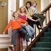 Four people sit on a staircase railing, smiling and posing together in a casual indoor setting with columns and a clock on the wall.
