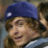 A pixelated close-up of a person wearing a blue cap with a white circular detail, smiling with eyes partially closed against a blurred background.