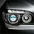 Pixelated close-up of a vehicle's front section showing two circular headlights with metallic silver trim and a dark background.