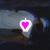 A pixelated heart-shaped icon with a bright pink center and white border, set against a dark, blurred background with gray and brown tones.