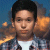 Pixelated portrait of a person with short dark hair wearing a white shirt under a dark patterned jacket against a blue background with orange shapes.