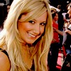 A static image of a person with long blonde hair wearing a black sleeveless top, smiling with visible teeth against a blurred background.