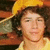 A person wearing a yellow cap and shirt with curly hair, shown in three variations of facial expression against a blurred background.