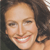 A static pixelated close-up of a smiling person with dark curly hair, light skin, and visible teeth, cropped to show the upper face and shoulder.