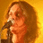 Pixelated image of a person with long brown hair holding a microphone against a yellow-orange gradient background.