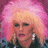 Pixelated image of a person with voluminous pink hair, light skin tone, and dark eyes, wearing large white earrings against a gradient background.