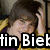A person with brown hair wearing a yellow jacket over a dark purple shirt, smiling with one hand on their head, partially obscured by text.