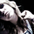 Pixelated close-up of a person's face with light-colored hair, dark clothing, and visible hand gesture against black background.