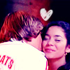 Two people embrace closely; one wears a white shirt with red text, the other has dark hair, and a white heart symbol floats above them.