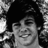 Black-and-white pixelated image of a person with dark hair, smiling face, and visible hand gesture against patterned background.