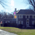 Pixelated image of a dark blue house with white trim, a chimney, and a flagpole holding a purple and yellow flag, set against a light blue sky with faint clouds and green grass in the foreground.