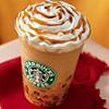 A close-up of a Starbucks iced coffee drink with whipped cream and caramel drizzle in a clear plastic cup on a red surface against an orange background.