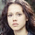 Pixelated close-up of a person with dark wavy hair, light skin tone, and neutral expression against blurred background.