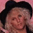 Pixelated image of a person with light skin tone wearing dark hat and long hair against pink background.