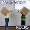 Two people stand indoors holding large brown cardboard boxes over their heads, reaching toward each other with outstretched arms as if to touch hands, with 'IMAGINATION' text at top and 'ROCKS' text at bottom.