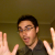 Pixelated image of a person with dark hair wearing glasses and a dark shirt, hands raised in front of a muted brown background.