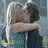 Two people embrace in a close-up pose with one person's arm around the other, set against an outdoor background.