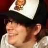 Person wearing black-and-white trucker hat with cartoon character on front, smiling, light-colored shirt, brown hair, dark background.
