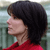 Pixelated profile view of a person with short dark hair wearing a red top against a blurred greenish background.