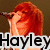 Pixelated image of a person with long red hair holding a microphone, overlaid with the text 'Hayley' in bold black letters on white background.