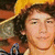 A person with curly brown hair wears a yellow hat and blue-and-white striped shirt against an orange background.