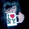 A person with dark hair holds a white mug with 'I ❤️ NY' text against a dark background.