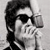 Black-and-white pixelated image of a person with dark hair wearing dark sunglasses, holding a microphone near their mouth against a light background.